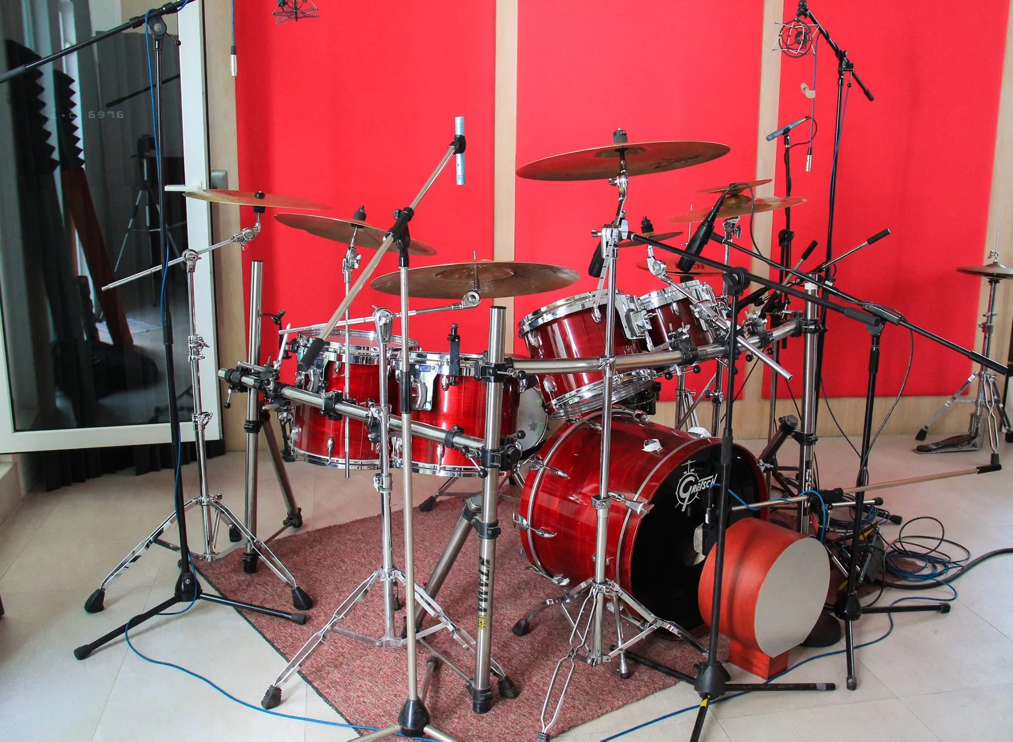 Batería acústica marca Gretsch de color rojo brillante armada en un estudio de grabación. El set incluye un bombo, tarola, múltiples toms y platillos montados sobre herrajes cromados. Toda la batería está rodeada por pedestales y micrófonos colocados estratégicamente para la captura de audio. Al fondo se aprecian paneles acústicos de color rojo brillante y beige sobre la pared.